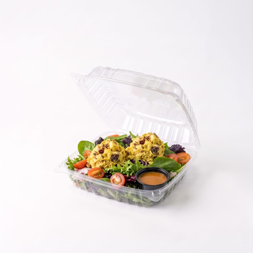 Open clear container holding leafy green salad with cherry tomatoes, creamy chicken salad, and a cup of dressing.