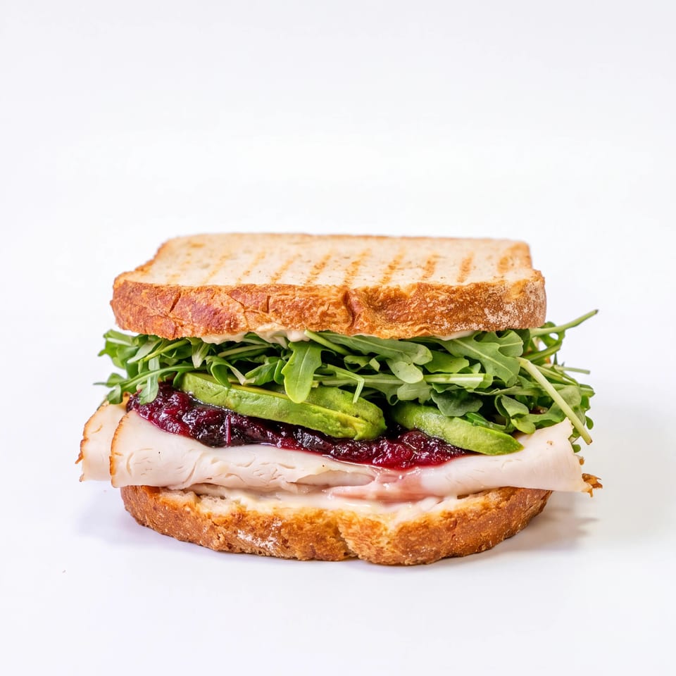 Grilled sandwich layered with turkey, cranberry sauce, avocado, arugula, and creamy spread on toasted bread.