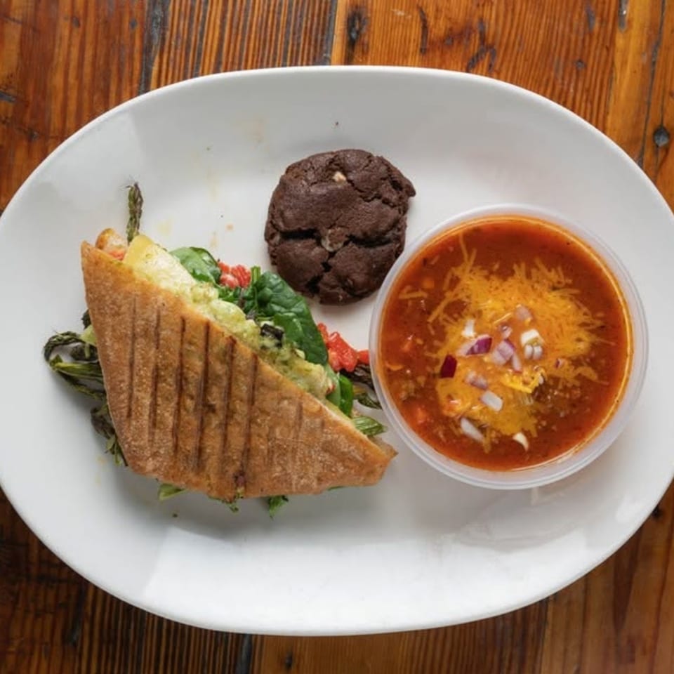 Plate with a grilled folded tortilla filled with greens, a bowl of reddish soup, and a chocolate cookie on a wooden table.