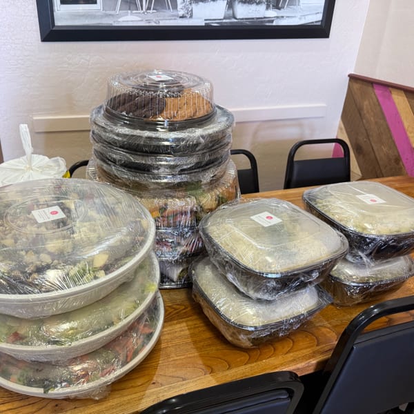 Multiple catering containers of food, including cakes and salads, on a wooden table at Bertha's Cafe.