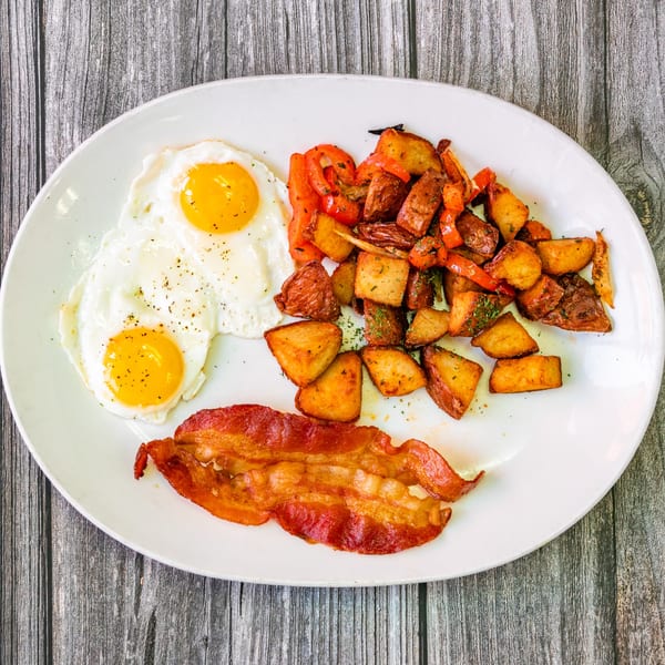 A Bertha's Cafe breakfast plate with two fried eggs, crispy bacon, and seasoned potatoes with peppers.