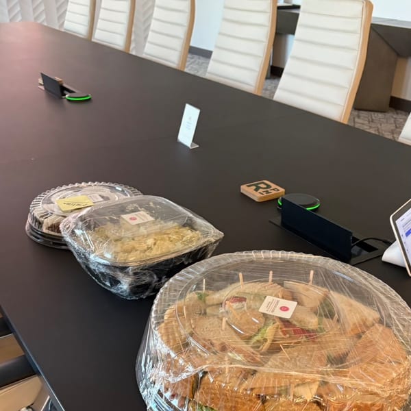 Catering containers of sandwiches and salads on a long black conference table with white chairs.