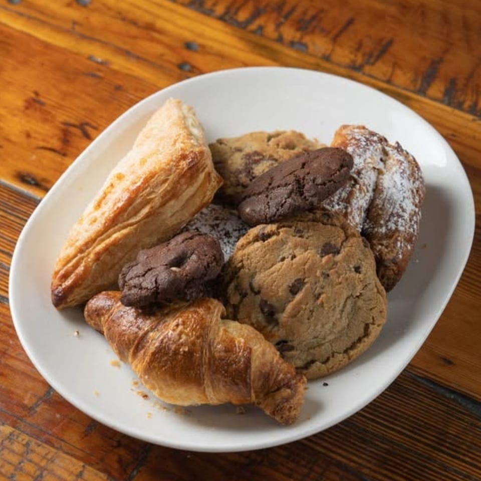 Assorted croissants and chocolate chip cookies arranged on a white plate atop a rustic wooden table.