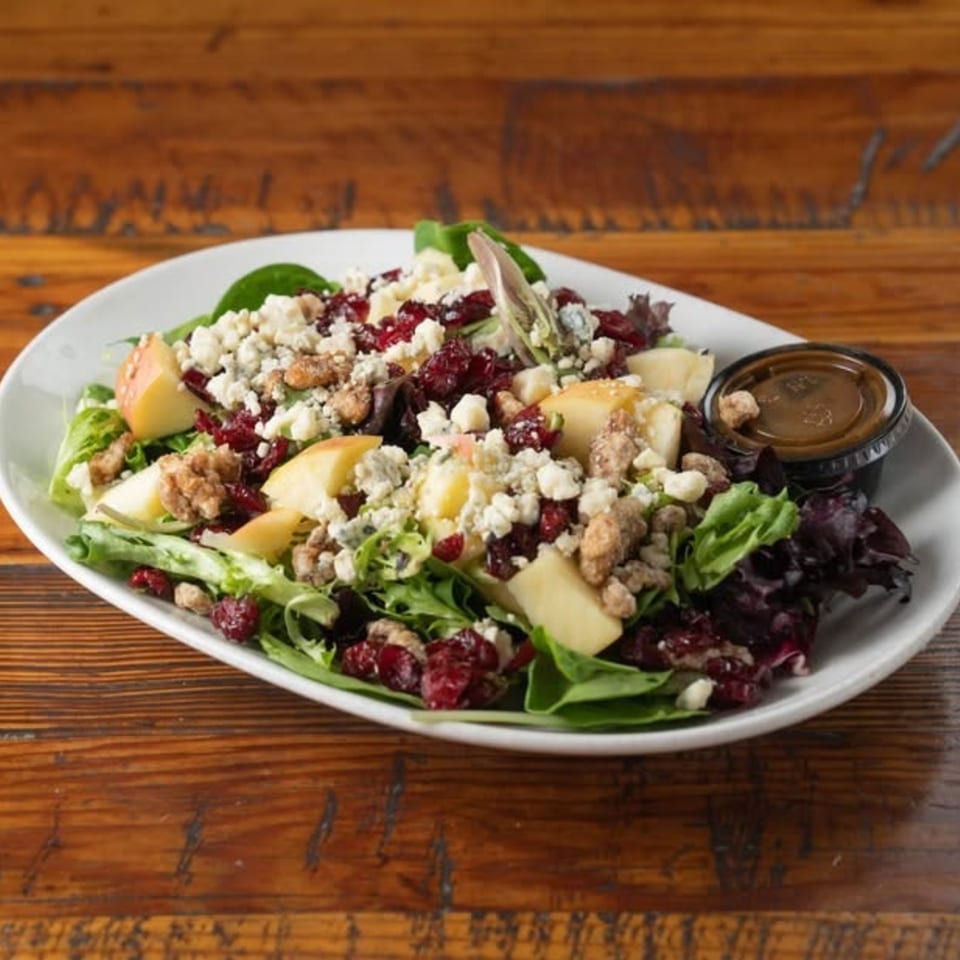 Mixed green salad with apple slices, dried cranberries, blue cheese, candied walnuts, and dressing on a white plate.