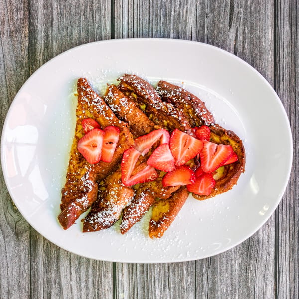 Golden French toast slices, dusted with powdered sugar and topped with fresh strawberries, on a white plate.
