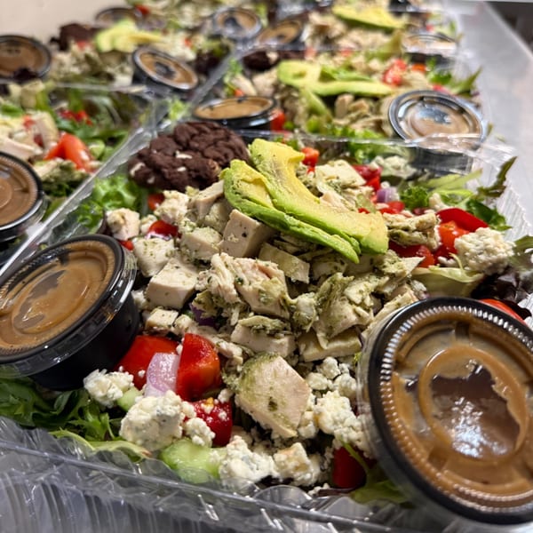 Multiple Bertha's Cafe salads in plastic containers, featuring chicken, avocado, tomatoes, and dressing.